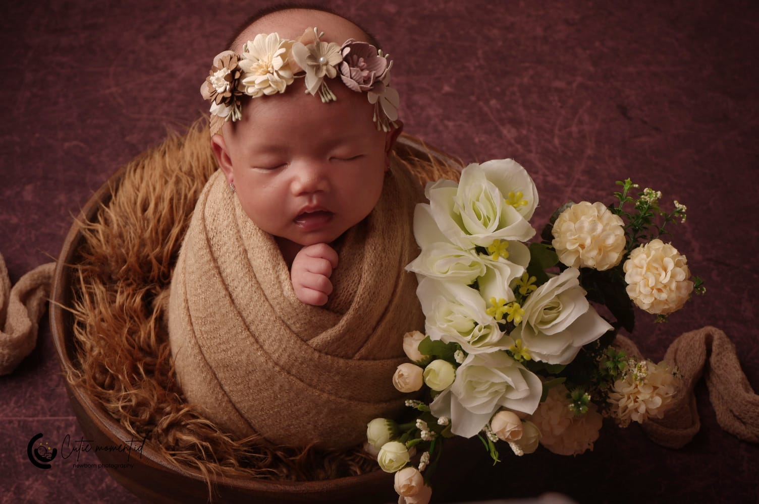 Foto bayi dibedong kain coklat di dalam mangkuk dengan hiasan bunga putih